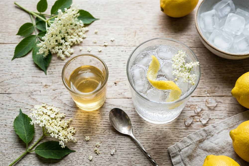 Measuring pale golden elderflower cordial beside a tall glass of sparkling water, lemon peel, and ice